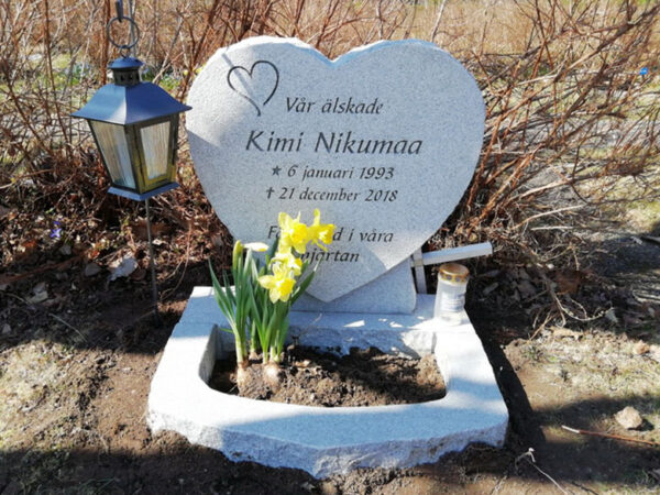 E019 i ljusgrå granit med finhuggen framsida och råarbetade sidytor. Gravyren är djupblästrad och lackad. Blomlist i oregelbunden form monterad framför sockeln.