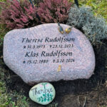 E048 i flammad indisk röd granit. Gravyren är försänkt svartmålad, bronssparvar på toppen.