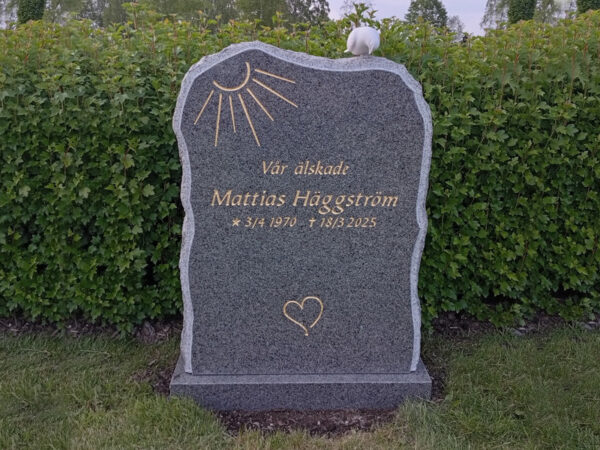 EN015 i polerad grå Bohusgranit, finhugget kantslag och råarbetade sidor. Polerad sockel, marmorduva, försänkt förgylld gravyr.